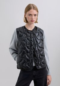 Black quilted vest with a glossy finish, round neckline, front zipper closure, worn over a light gray long-sleeve top, with textured sleeves.