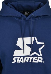 Kék színű kapucnis pulóver, pamut keverékből készült, nagy fehér logóval, amely csillagot és a márkanevet tartalmazza. Kihúzható kapucnival és puha tapintással rendelkezik.