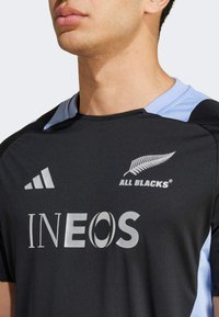 Černý atletický dres s světle modrými akcenty, zdobený stříbrnými logy: "ALL BLACKS" a "INEOS," vyrobený z hladké, syntetické látky.