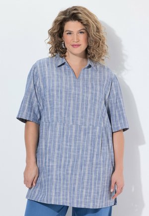 Frau mit lockigem Haar, die eine blaue, gestreifte Tunika mit kurzen Ärmeln und blaue Hosen trägt, steht vor einem weißen Hintergrund.