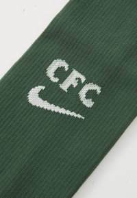 Grüne Sportsocke mit weißen "CFC"-Text und Nike-Logo, flach auf einem weißen Hintergrund ausgelegt.