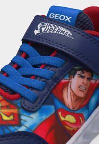 Κοντινό πλάνο μπλε αθλητικού παπουτσιού με γραφικό του Σούπερμαν, μπλε κορδόνια, κόκκινα διακοσμητικά στοιχεία και μπλε λουράκι με την επιγραφή "Superman" και μπλε γλώσσα με την επιγραφή "GEOX".