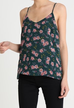Vrouw die een donkergroene mouwloze top met bloemenprint en dunne bandjes draagt, gecombineerd met een zwarte broek tegen een effen achtergrond.