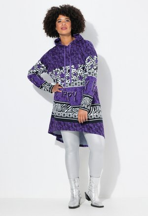 Kvinde med krøllet hår iført en lilla mønstret hættetrøjekjole, sølvleggings og sølv ankelstøvler, stående mod en hvid baggrund.