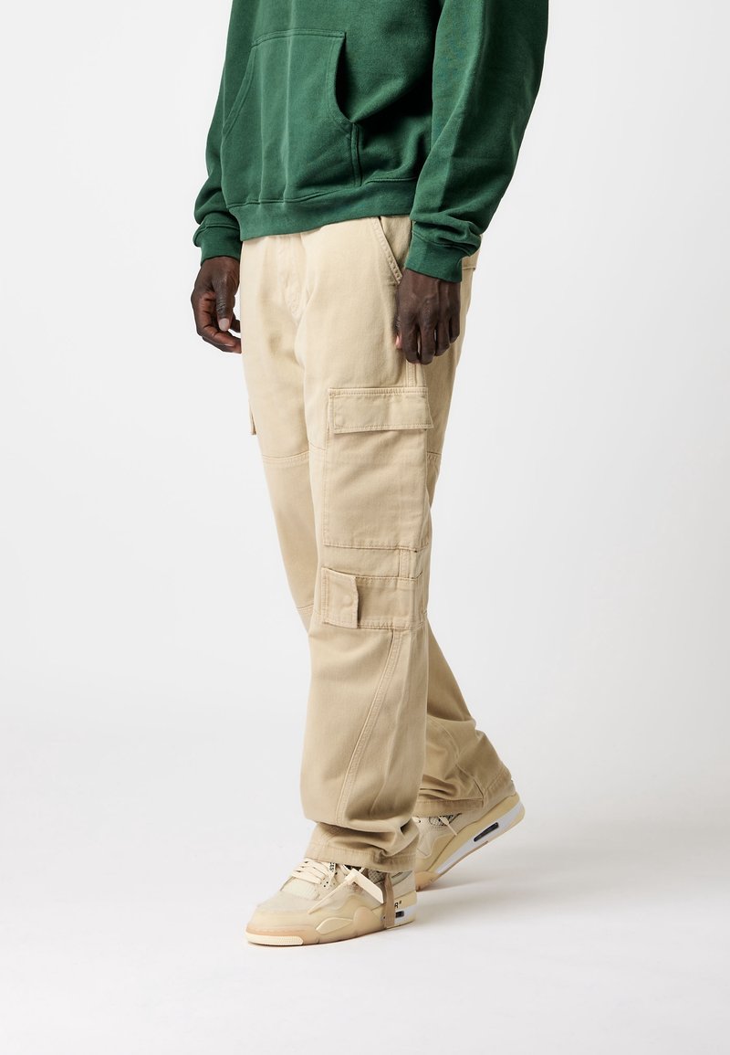 Pantalones cargo beige con múltiples bolsillos, corte cómodo y tejido resistente, combinados con zapatillas beige y una sudadera con capucha verde.