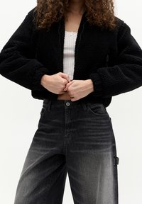 Black cropped bomber jacket made of fluffy fleece, featuring a zip closure and voluminous sleeves, paired with high-waisted faded black jeans.