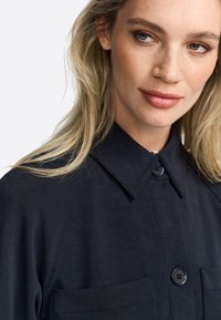 Chemise noire avec un col, deux poches avant, et fermeture à boutons. Texture lisse avec des détails de couture subtils et coupe ajustée.