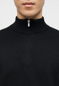 Άνδρας που φοράει μαύρο ριμπ πλεκτό πουλόβερ με ψεύτικο λαιμό και ασημένιο φερμουάρ κλειστό στον γιακά, εμφανίζοντας μόνο το κάτω μέρος του προσώπου και τον λαιμό.