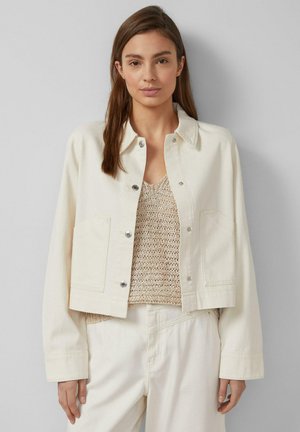 Kobieta o prostych brązowych włosach nosi kremową krótką kurtkę na beżowym dzianinowym topie i dopasowanych kremowych spodniach, stoi na tle szarej ściany.