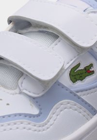 Λευκό αθλητικό παπούτσι με διπλά λουριά βέλκρο και πράσινο λογότυπο κροκόδειλου στο πλάι, με ανοιχτό μπλε και γκρι λεπτομέρειες.