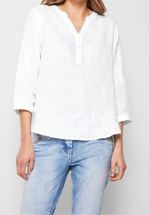 Kobieta ubrana w białą lnianą koszulę z długim rękawem i dekoltem w serek oraz jasnoniebieskie wyblakłe dżinsy, stoi z ramionami swobodnie opuszczonymi wzdłuż ciała.