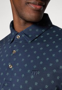 Polo navy con cuello que presenta un patrón floral verde, pequeños botones y un logo bordado en el pecho.