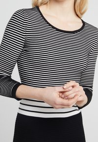 Femme portant un haut à manches longues noir et blanc à rayures horizontales, les mains jointes devant au niveau de la taille.