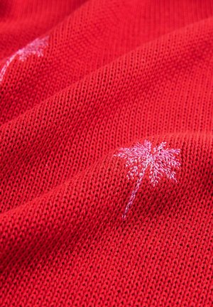 A piros kötött anyagnak sima textúrája van, amelyet világos rózsaszín pálmafák hímzése díszít, ezzel dekoratív elemet adva hozzá.