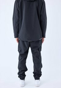 Chaqueta impermeable negra con capucha y un ajuste oversize, combinada con pantalones cargo negros que tienen bolsillos laterales y puños elásticos. Zapatillas blancas completan el look.