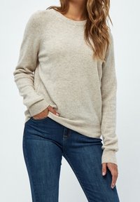 Kvinna som bär en beige långärmad tröja och mörkblå jeans, med en hand i fickan och vågigt hår över axeln.
