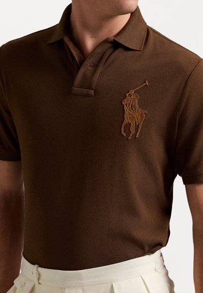 Cămașă polo maro dintr-un material texturat; are un logo brodat cu un călăreț, în nuanță mai deschisă, pe partea stângă a pieptului.