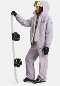 Snowboarder in een lichtpaarse waterdichte jas en broek, houdt een snowboard vast. Heeft zwarte handschoenen en een helm met skibril.