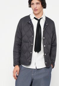 Quiltet mørkegrå jakke med trykknapper, med en rund krage, båret over en hvit skjorte og svart slips, kombinert med blå bukser.