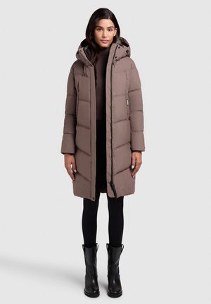 Doudoune marron avec capuche, dotée d'une fermeture éclair et d'un design matelassé. Portée par-dessus un haut sombre et associée à des leggings noirs et des bottes.