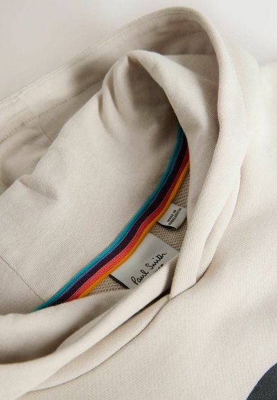 Col col de vêtement beige avec bordure rayée arc-en-ciel et étiquette de marque "Paul Smith", montrant le tissu plié et les détails de couture.