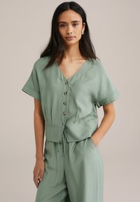 Blusa verde chiaro a maniche corte con scollatura a V, chiusura a bottoni e vita regolabile con coulisse. Tessuto morbido e vestibilità comoda, abbinata a pantaloni coordinati.