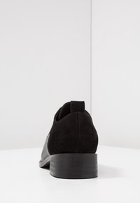 Chaussure en daim noir à faible talon, avec une finition lisse et un design minimaliste. La chaussure a un bout arrondi et une languette à l'arrière.