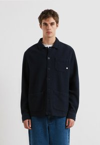 Junger Mann, der eine dunkelblaue, geknöpfte Strickjacke mit Taschen vorne und Kragen trägt, kombiniert mit blauen Jeans, vor einem schlichten Hintergrund stehend.