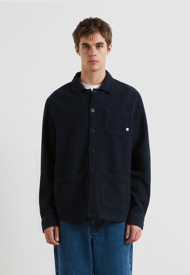 Junger Mann, der eine dunkelblaue, geknöpfte Strickjacke mit Taschen vorne und Kragen trägt, kombiniert mit blauen Jeans, vor einem schlichten Hintergrund stehend.