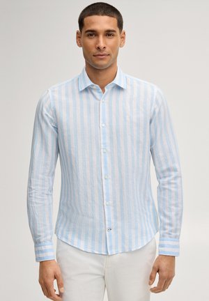 Mann trägt ein hellblau-weiß gestreiftes, langärmliges Hemd mit Knöpfen und weiße Hose, steht vor einem schlichten Hintergrund.