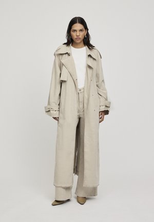 Long manteau beige avec un grand col, devant croisé à double boutonnage et poignets retroussés, associé à un pantalon taille haute assorti et des chaussures à bout pointu.