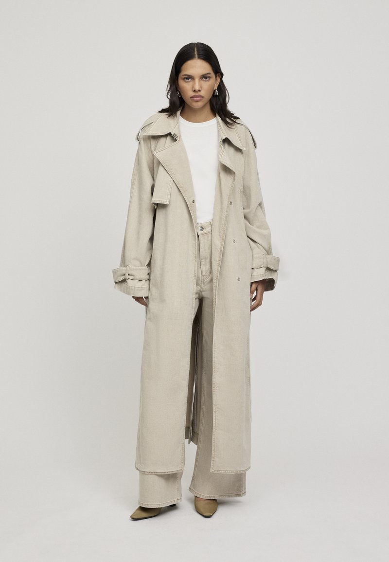 Lungo trench coat beige con colletto ampio, frontale doppiopetto e polsini arrotolati, abbinato a pantaloni a vita alta coordinati e scarpe a punta.
