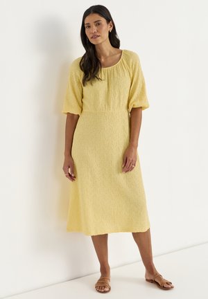 Kvinde med langt mørkt hår iført en tekstureret gul midi-kjole og brune flade sandaler, stående op ad en hvid væg.