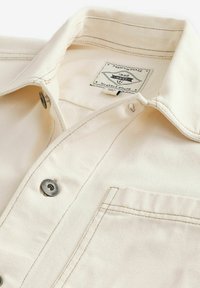 Camisa de mezclilla color crema con cuello puntiagudo, cierre de botones plateados y un bolsillo en el pecho. Presenta una etiqueta de tela en el interior del cuello.