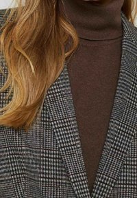 Bleiseri mustavalkoisessa houndstooth-kuviossa ruskean poolopaidan päällä, jossa on särmikkäät kaulukset ja teksturoidut kankaat.