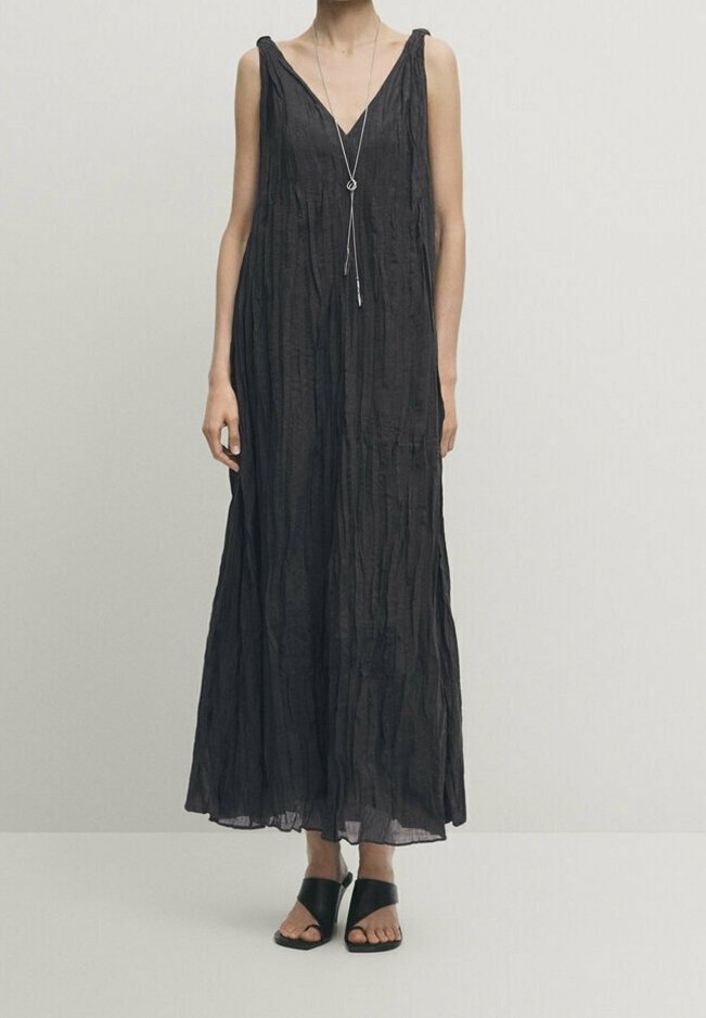 Robe maxi noire sans manches en tissu froissé avec un décolleté profond en V, portée avec des sandales à talons noires ouvertes et un long collier argenté.