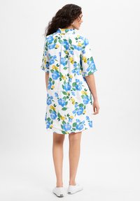 Weißes Kleid mit blauen Blumen- und gelben Zitronenmuster, kurzen Ärmeln, einem Kragen und seitlichen Schlitzen, kombiniert mit weißen Sneakers.