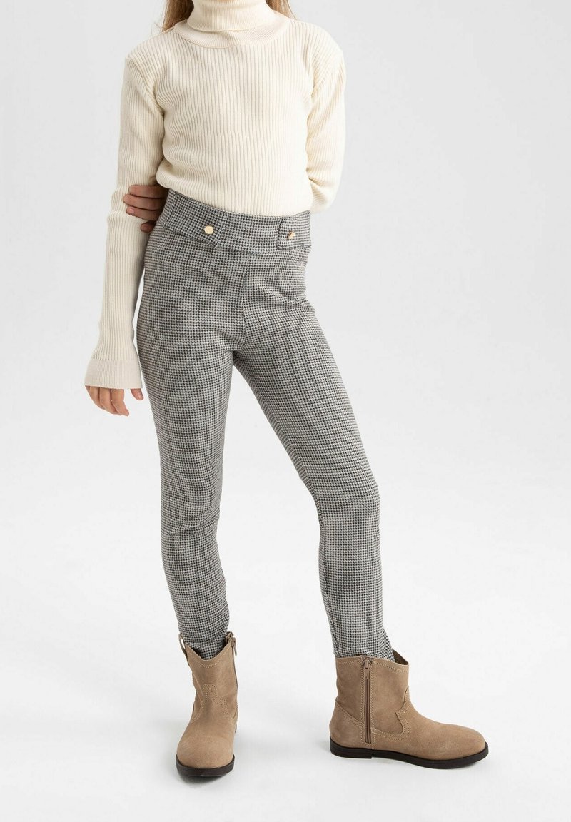 Enfant portant un col roulé côtelé crème, un pantalon gris à taille haute motif pied-de-poule avec des boutons dorés, et des bottines en daim beige, debout sur fond blanc.