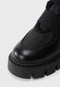 Fekete bokacsizma sima bőr felsőrésszel és puha szövet részletekkel. Jellemzője a vastag gumitalp, amely mély barázdákkal rendelkezik a tapadás érdekében.