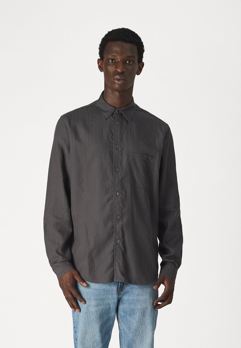 Mørkegrå langermet skjorte laget av et glatt stoff, med knappestolpe foran, krage og en liten brystlomme. Brukt med lyseblå jeans.