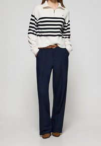 Femme portant un pull rayé blanc et bleu marine, un pantalon large bleu marine avec une ceinture marron, et des chaussures marron, debout les mains dans les poches.