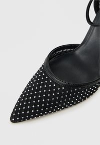 Zapato negro de punta afilada con parte superior de tela texturizada adornada con strass plateados en un patrón de cuadrícula, con un elegante ribete de cuero.
