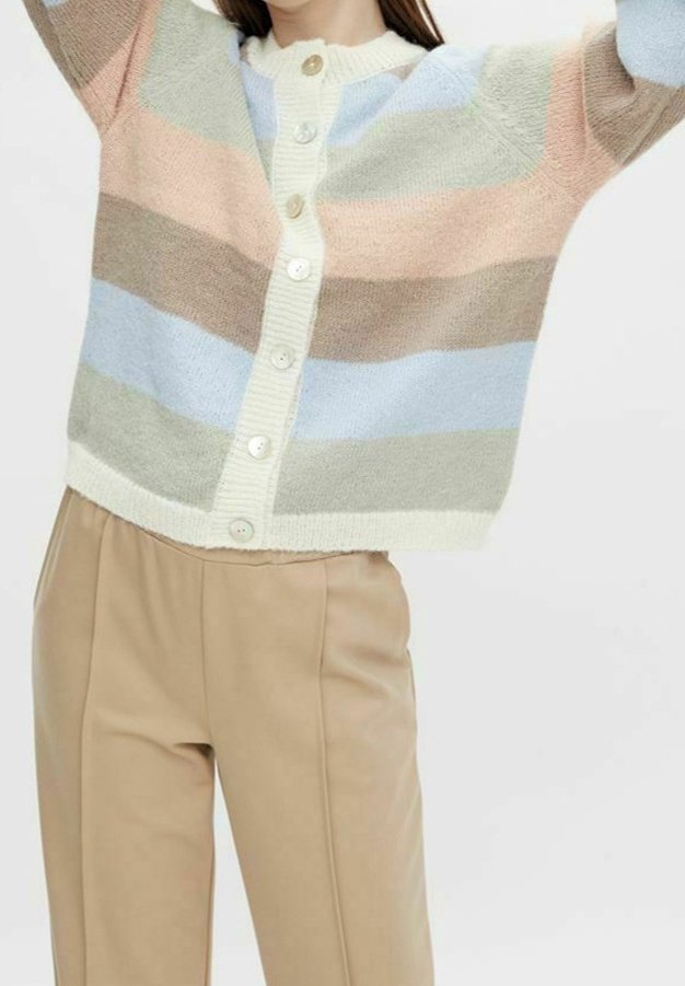 Personne portant un cardigan à rayures pastel avec de gros boutons et un pantalon beige taille haute, levant les bras au-dessus de la tête.