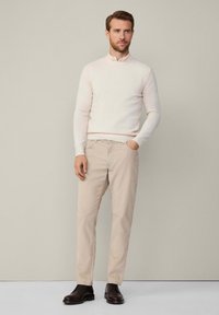 Světle béžový svetr s kulatým výstřihem, spárovaný se světle khaki chino kalhotami a tmavě hnědými botami. Hladká textura a dobře padnoucí design.