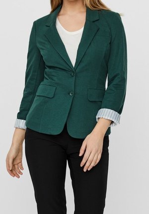 Mujer con blazer verde oscuro con puños a rayas remangados, camisa blanca y pantalones negros, mostrada desde los hombros hasta las caderas.