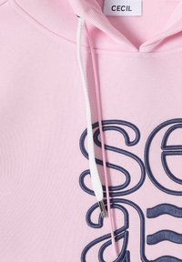 Sweat à capuche en coton rose avec texte en cursive bleu marine et cordons blancs ornés d'une pointe métallique. Étiquette visible au niveau de l'encolure.