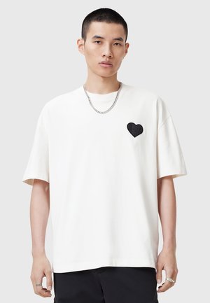 Hvid bomulds T-shirt med korte ærmer, der har et sort hjerte-design på brystet og en afslappet pasform. Båret med en sølv kæde halskæde.