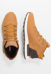 Scarpe da trekking in pelle marrone con un design alto, dettagli in mesh beige, lacci gialli, accessori in oro e una suola in gomma bianca e grigia.