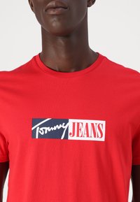 Rød bomulds t-shirt med et kontrasterende grafiktryk i marineblå og hvid af "Tommy" og "JEANS" på forsiden. Klassisk crew neck design.