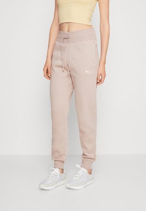Femme portant un pantalon de jogging beige avec cordon à la taille, des baskets blanches et un débardeur beige court, debout devant un fond uni.